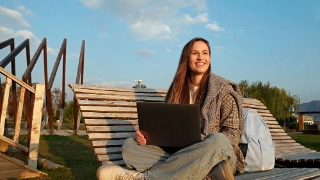 Eine junge Frau sitzt am einen sonnigen Tag auf einer Bank und arbeitet am Laptop.